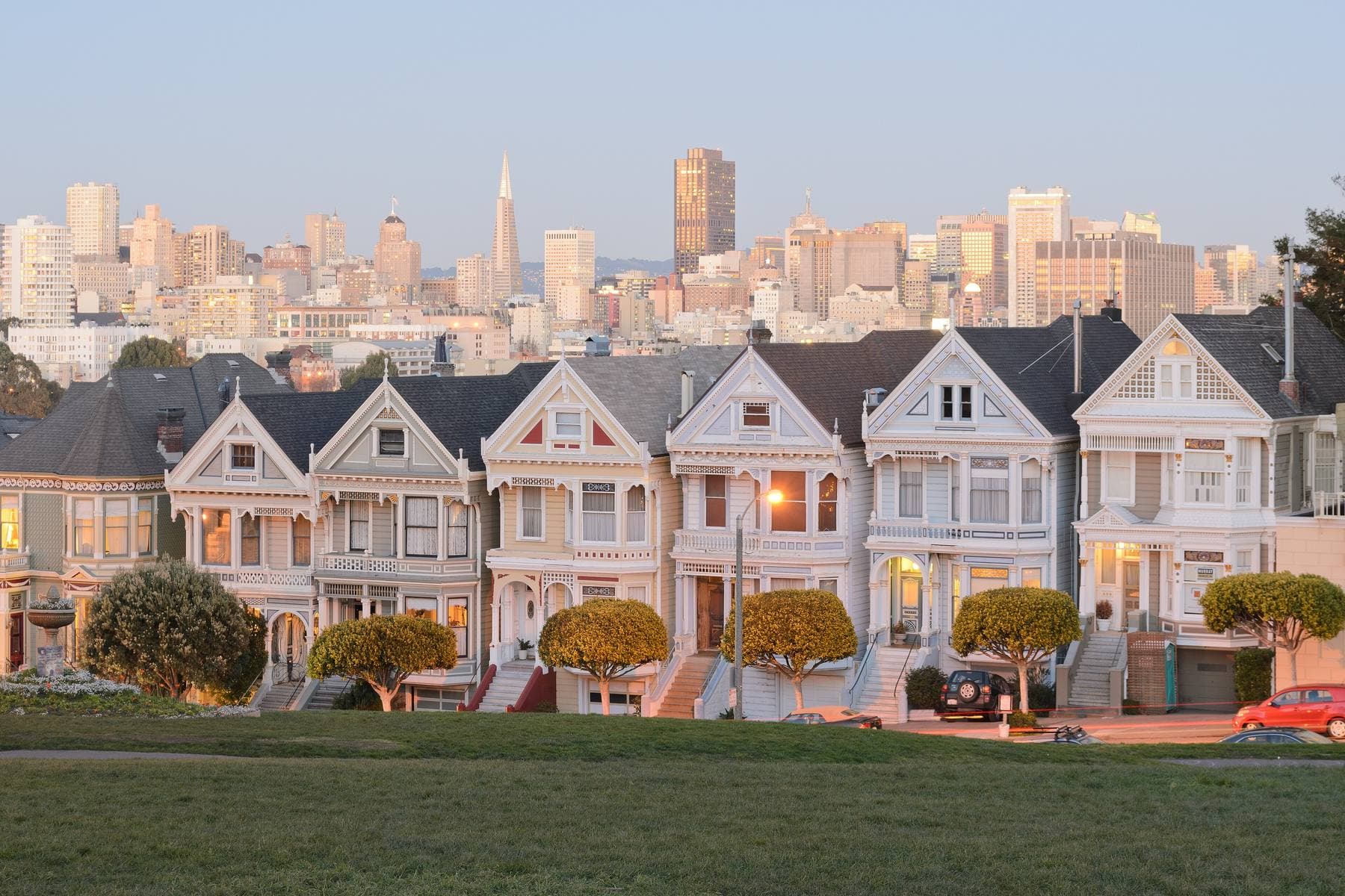 A row of Victorian houses facing Alamo Square at dusk, with lit windows and San Francisco's downtown skyline behind them
