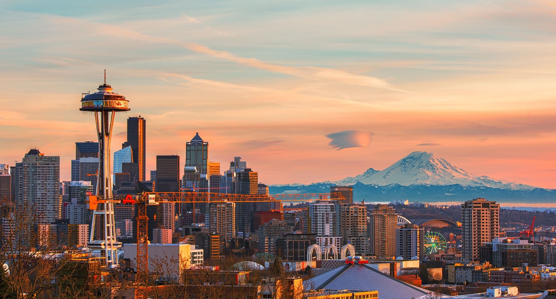 The Seattle skyline and Space Needle glowing at sunset, with snow-capped Mount Rainier on the horizon under a pink and orange sky