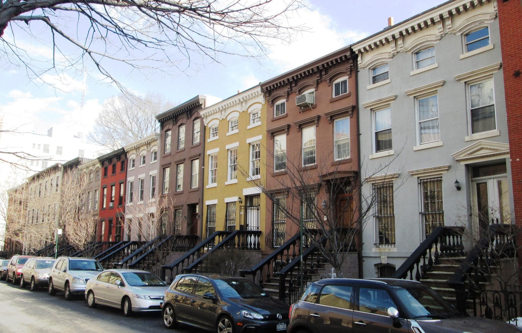 A sunlit row of colourful Italianate townhouses with black iron stoop railings and bare winter trees along St. Felix Street